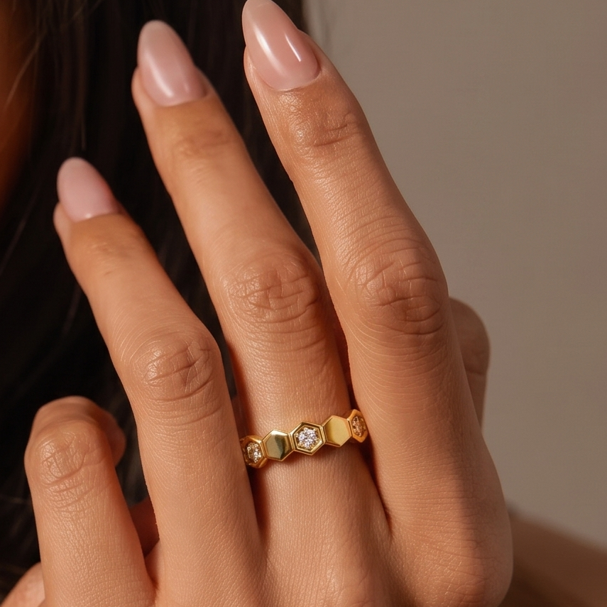 A close-up of a hand wearing a delicate gold ring with small green and clear gemstone accents, set against a soft neutral background.