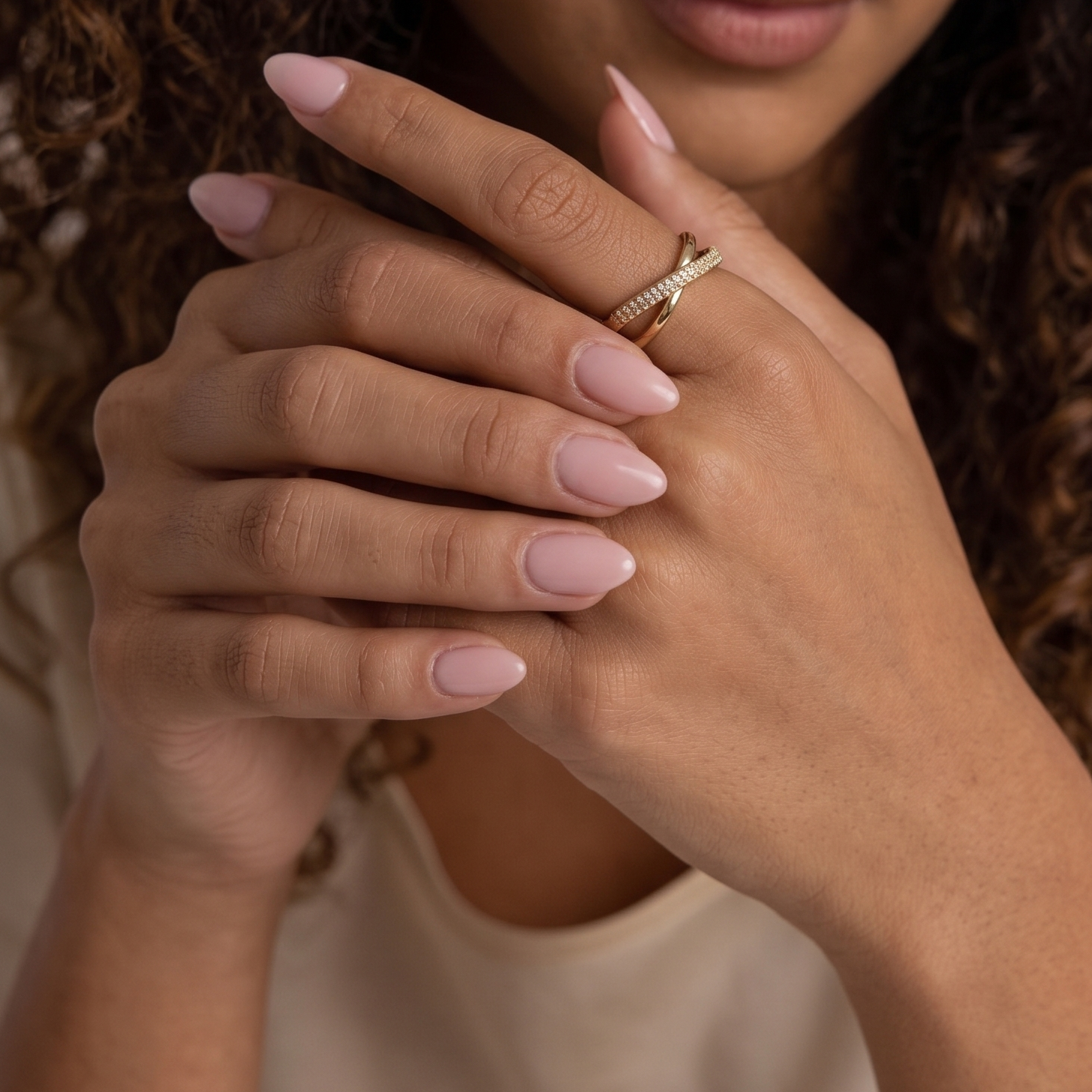 A close-up of a person's hands with long, pale pink manicured nails, wearing a delicate gold ring with small clear stones. The background is softly blurred, showing warm skin tones and dark curly hair.