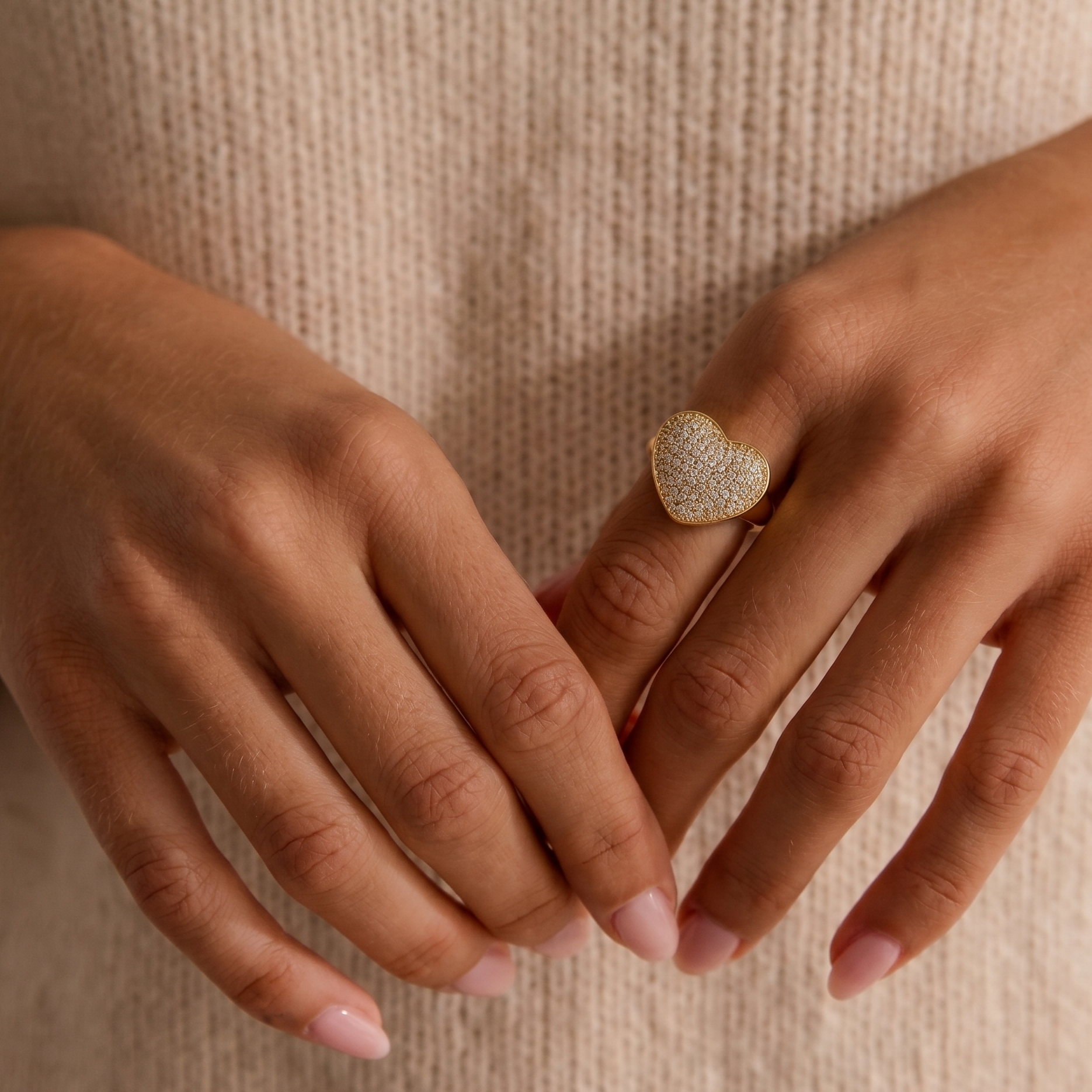 A close-up of a person's hands wearing a gold heart-shaped ring adorned with small clear stones, set against a soft beige knit sweater.