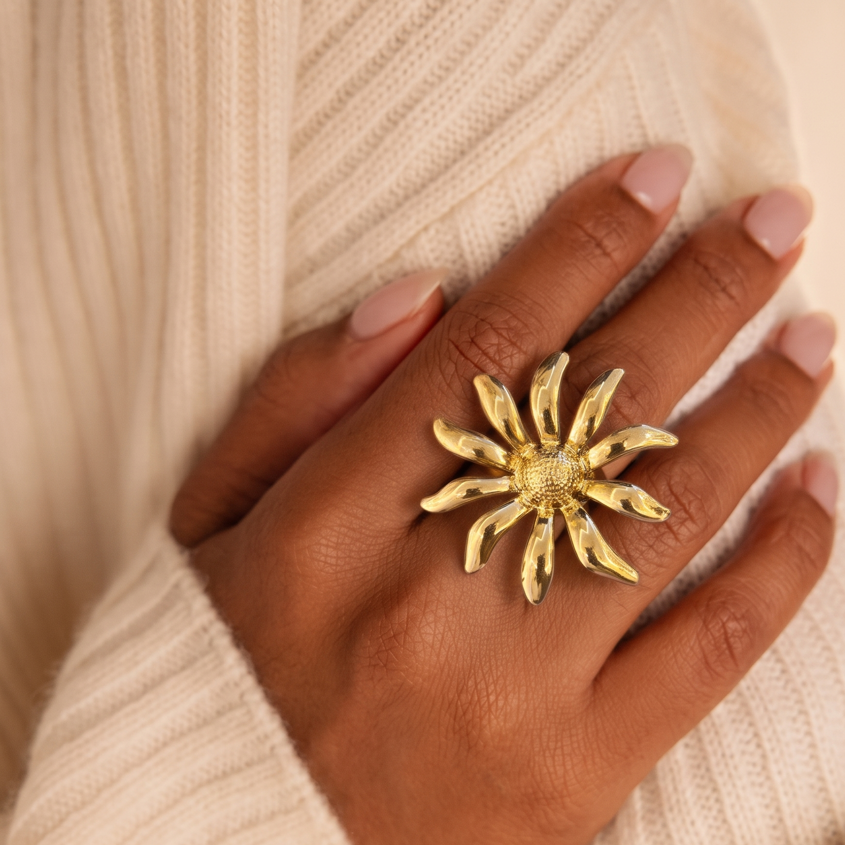 A close-up of a hand wearing a large, textured gold sunburst ring, styled with a cream-colored ribbed knit sweater.