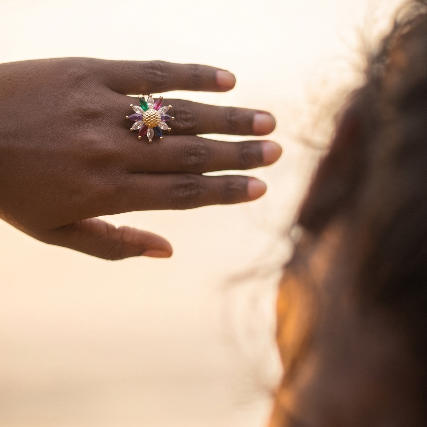 A close-up lifestyle shot of a person's hand wearing a colorful sunburst ring, with the back of their head and shoulder visible in warm, golden-hour light against a soft, blurred background.