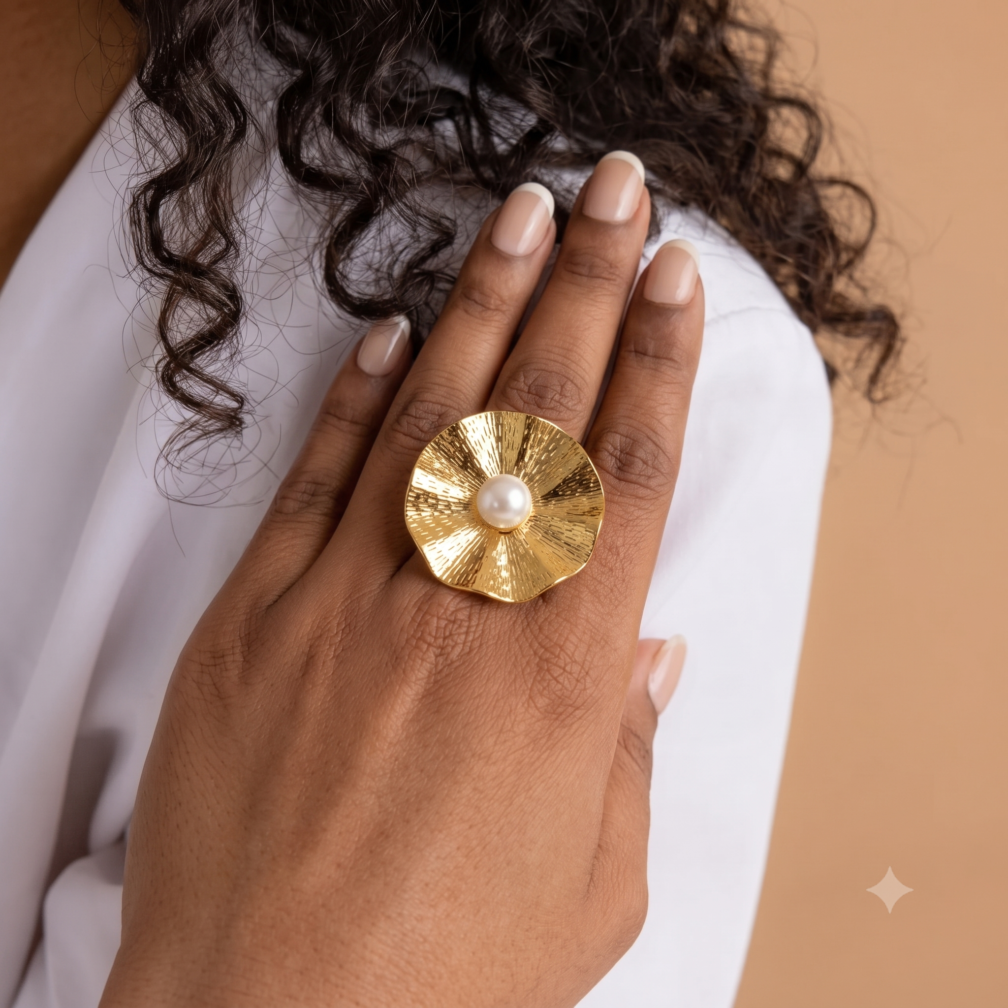 A close-up of a hand wearing a large gold ring with a textured, petal-like circular design and a central white pearl, set against a white top and warm beige background.
