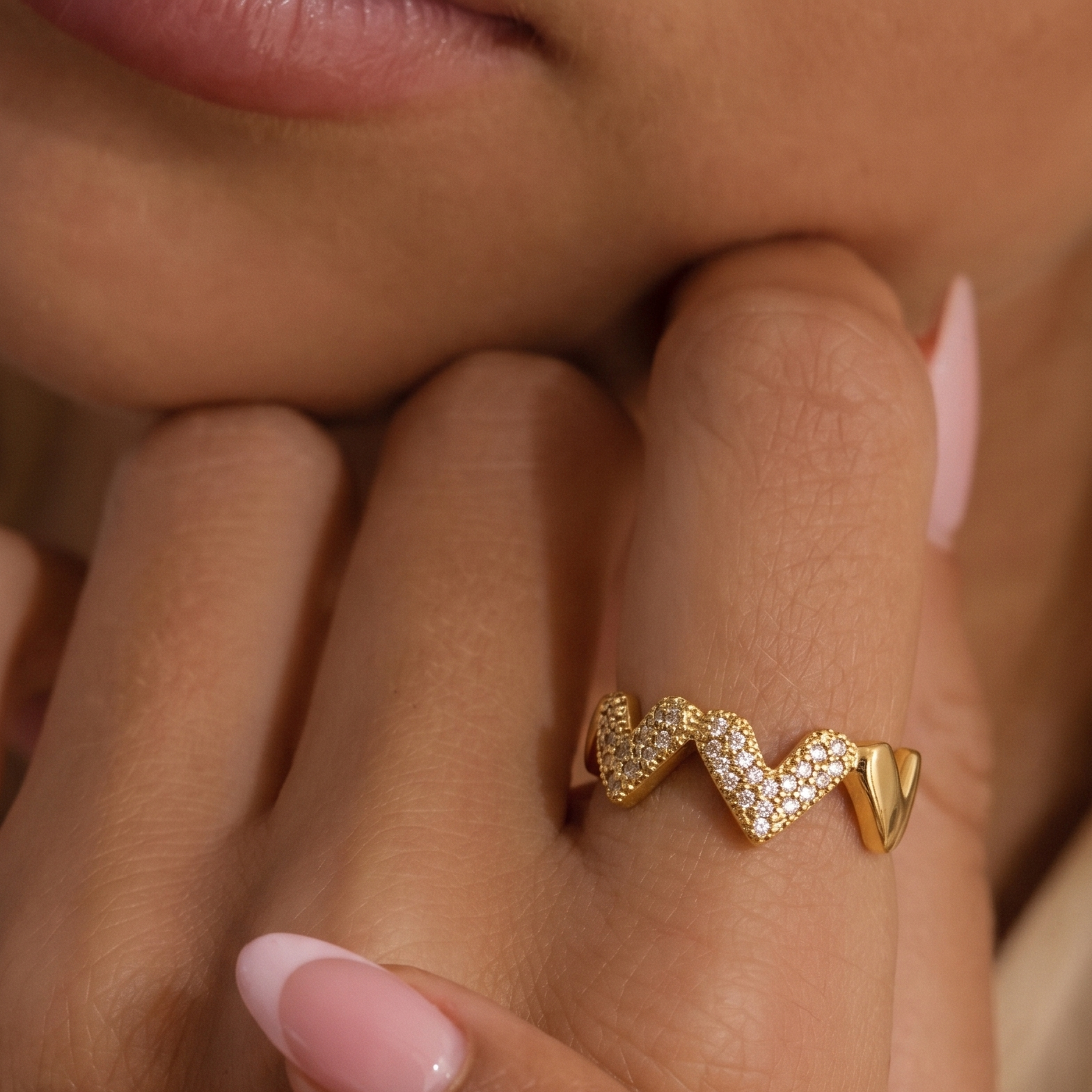 A gold ring with a wavy, textured pavé design worn on a finger, shown in a close-up lifestyle shot of a person's hand and lower face.