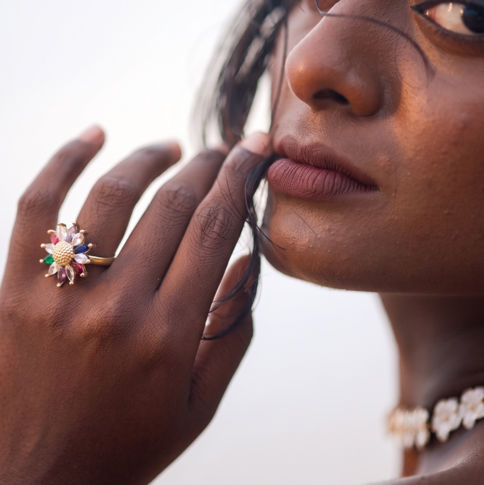 A close-up lifestyle shot of a model wearing a colorful floral ring on her finger and a white flower choker necklace against a bright, soft background.