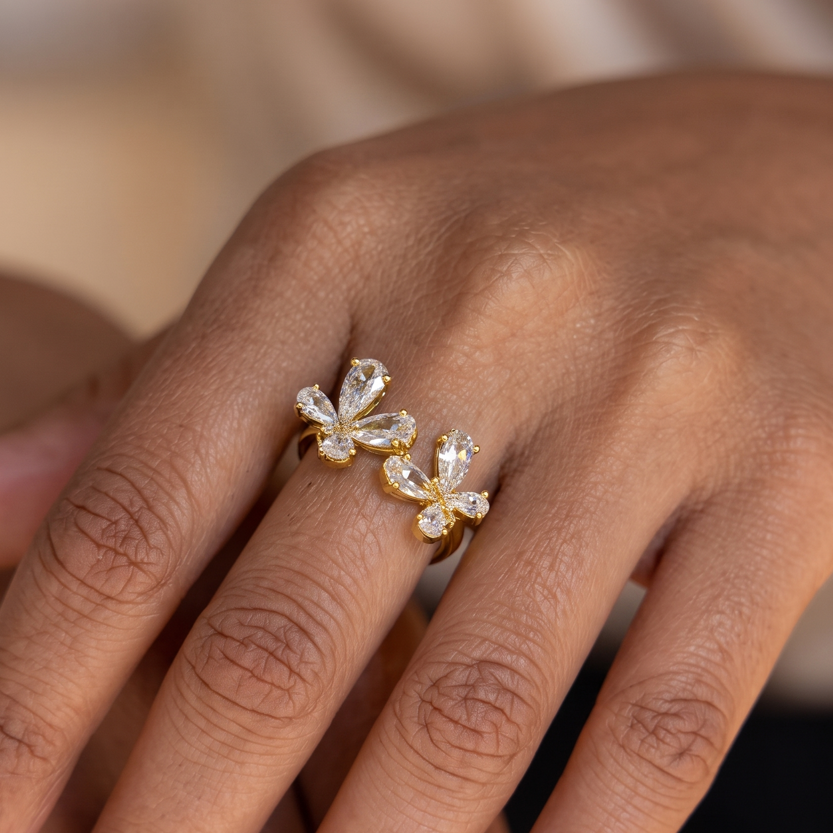 A close-up of a person's hand wearing a gold ring with two flower-shaped clear gemstone clusters.