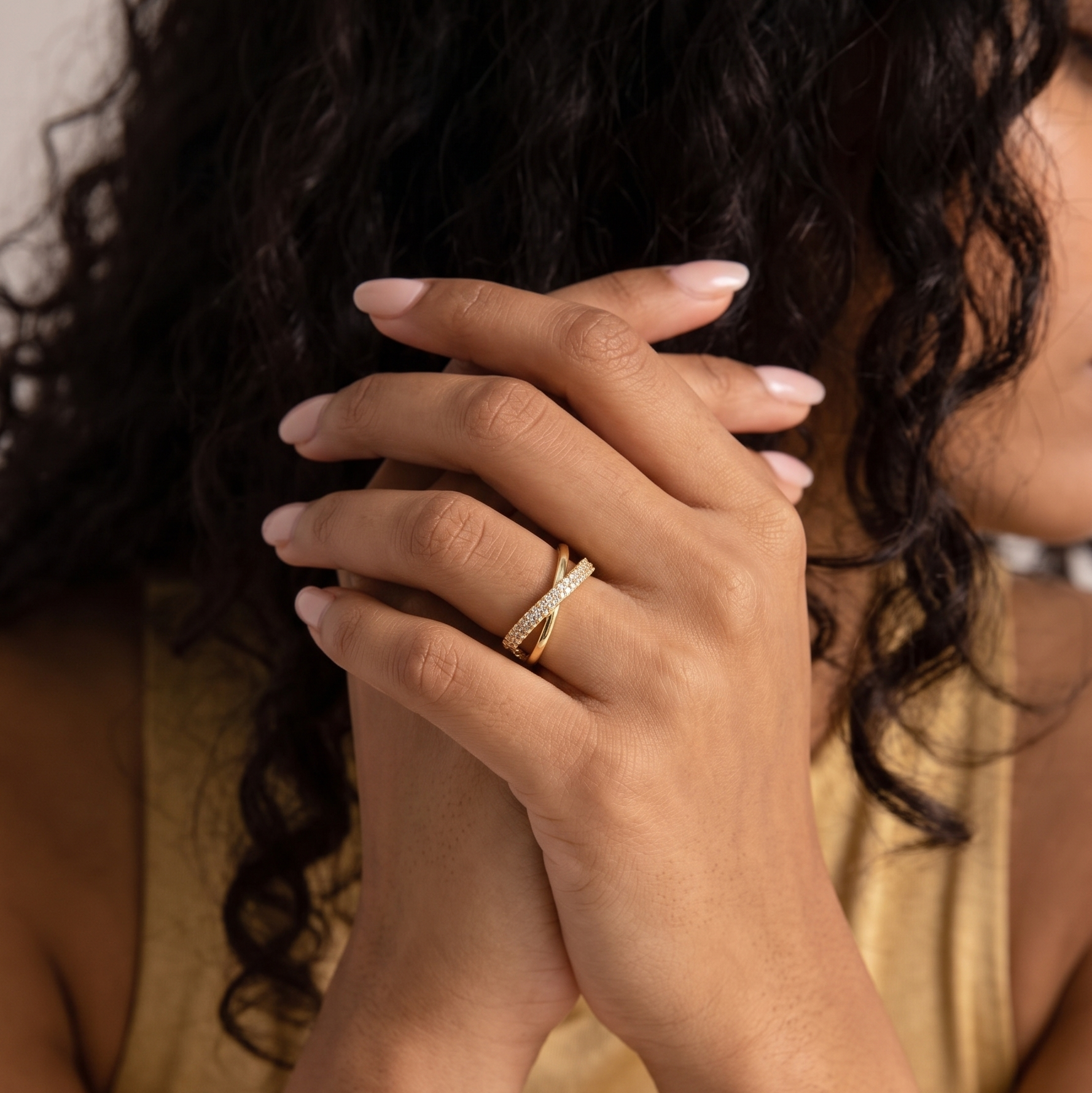 A close-up lifestyle shot of a woman with dark curly hair wearing a delicate gold ring with a twisted design and small clear stones on her finger, with her hands clasped in front.