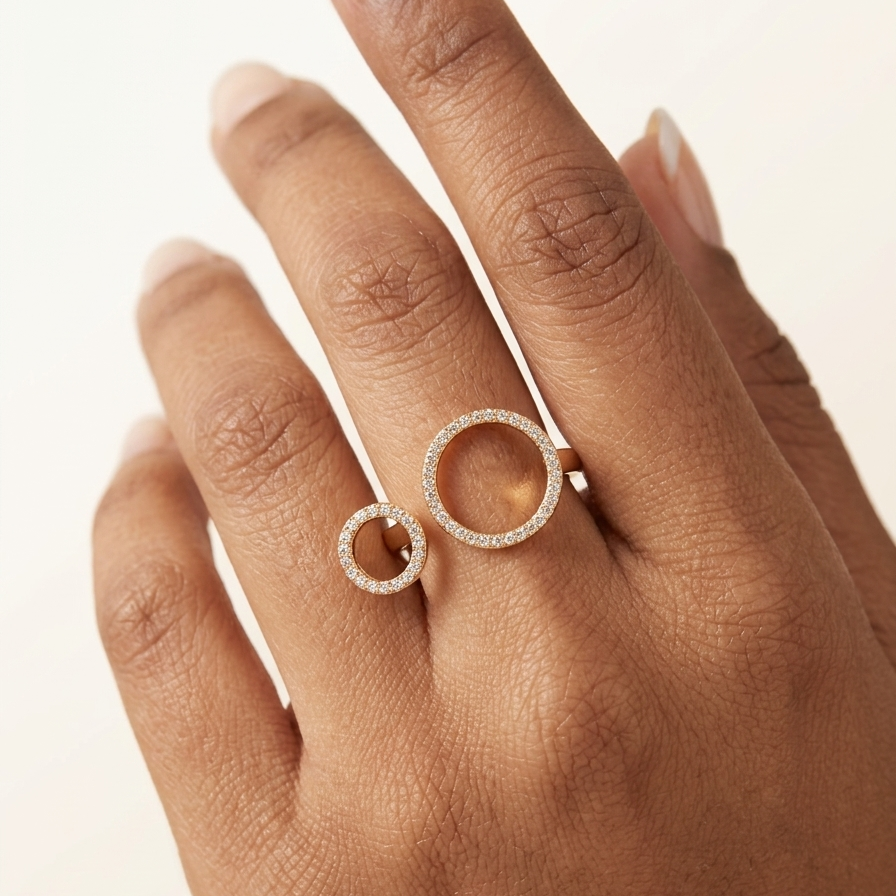 A close-up of a hand wearing two stacked silver rings, each featuring a textured, open circular design set with small clear stones, against a warm off-white background.