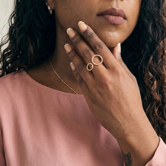A close-up lifestyle shot of a woman wearing a delicate gold ring with two interlocking oval loops on her finger, paired with a soft pink top.