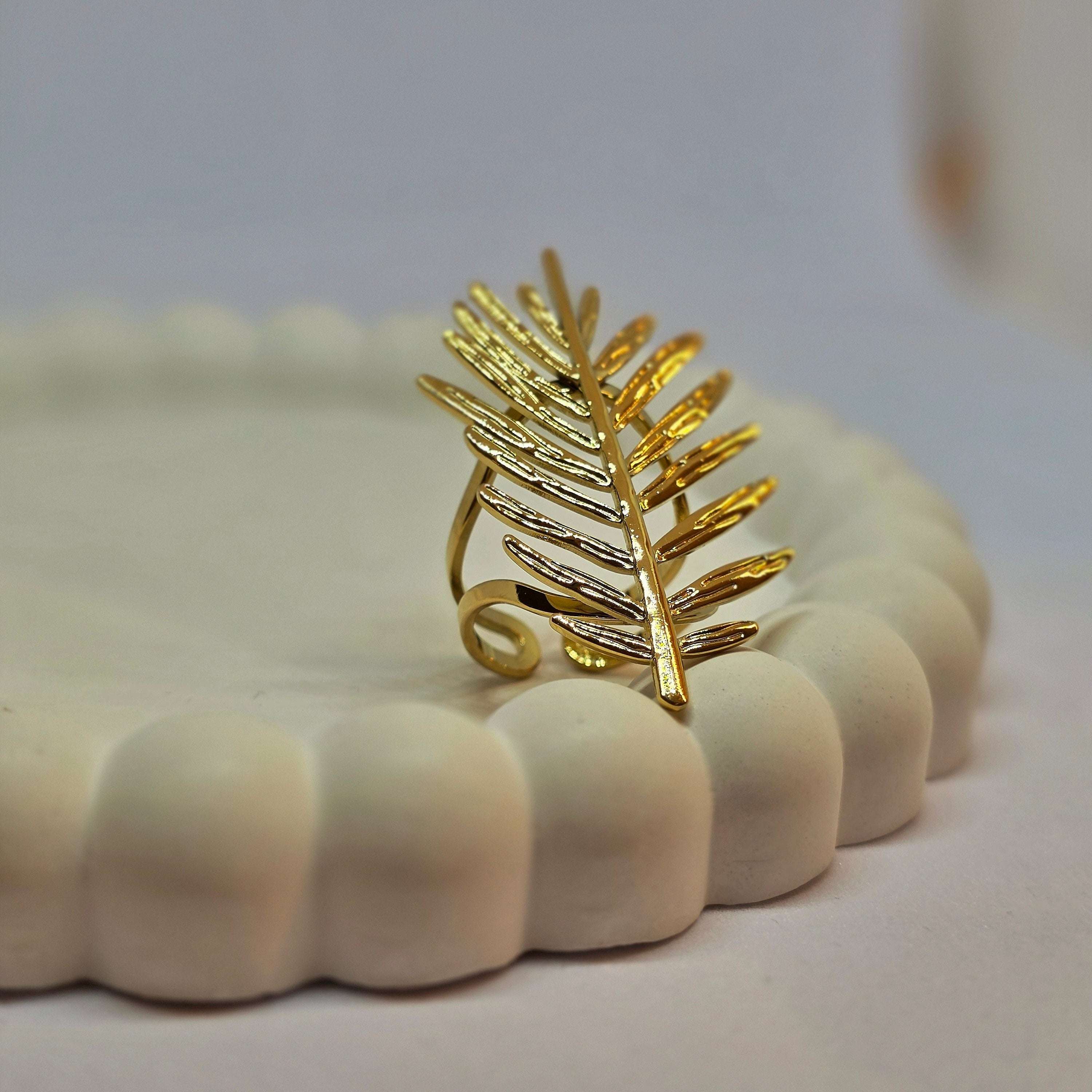 A gold-toned leaf-shaped ring displayed on a small white scalloped dish against a soft light gray background.