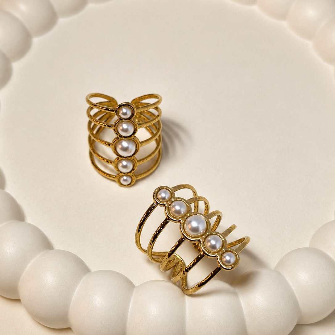 Two gold-plated multi-band rings with white faux pearls, displayed on a round white beaded tray against a warm cream background.