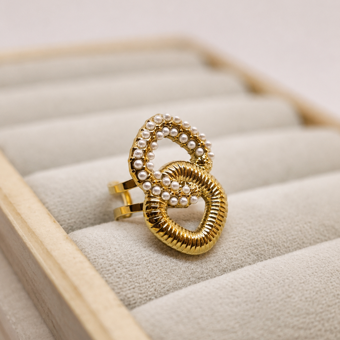 A gold ring with two interlocking textured loops, one adorned with small white pearls, displayed on a beige jewelry box cushion.
