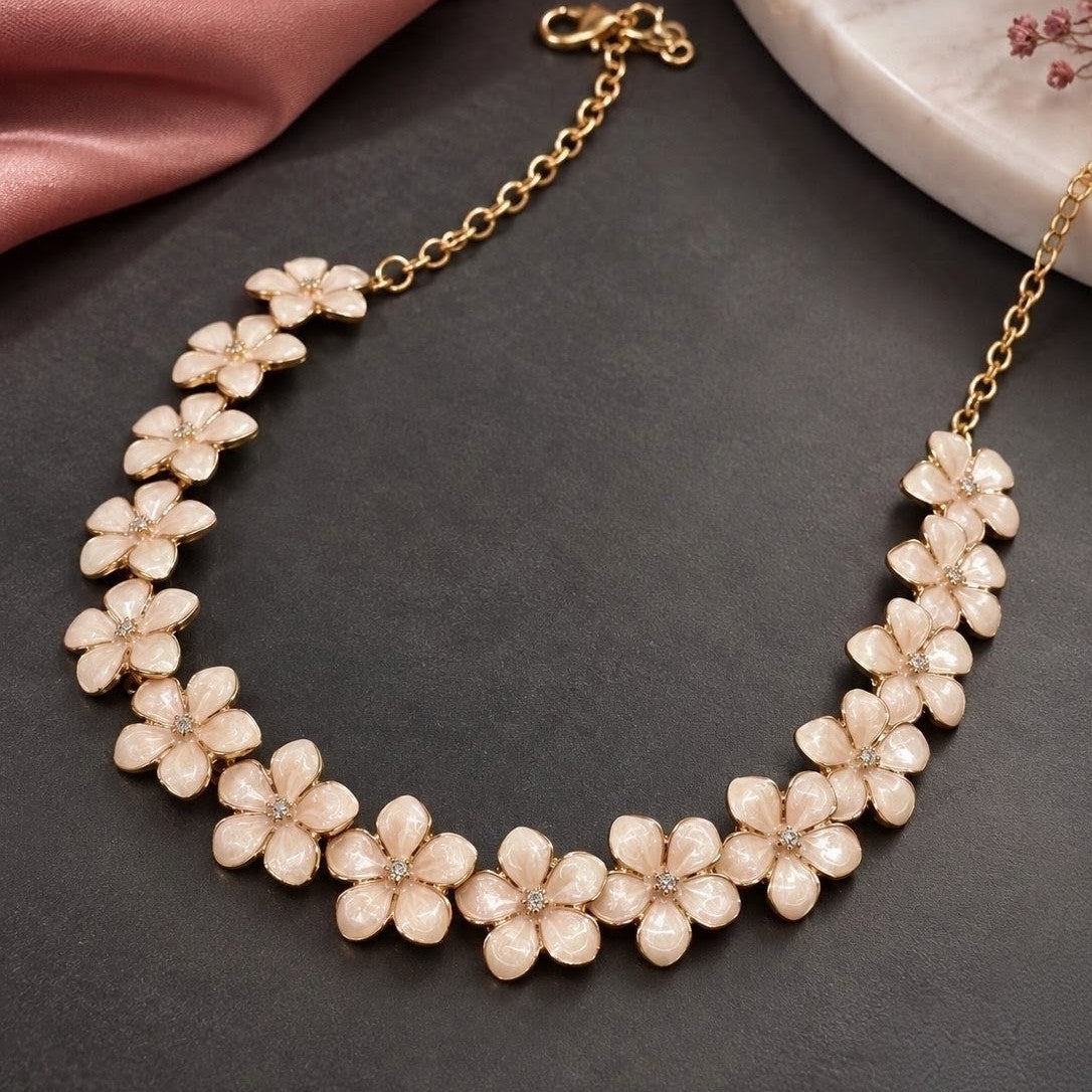 A gold-tone floral collar necklace with pale pink flower petals, displayed on a dark gray textured surface with pink satin and a marble plate in the background.