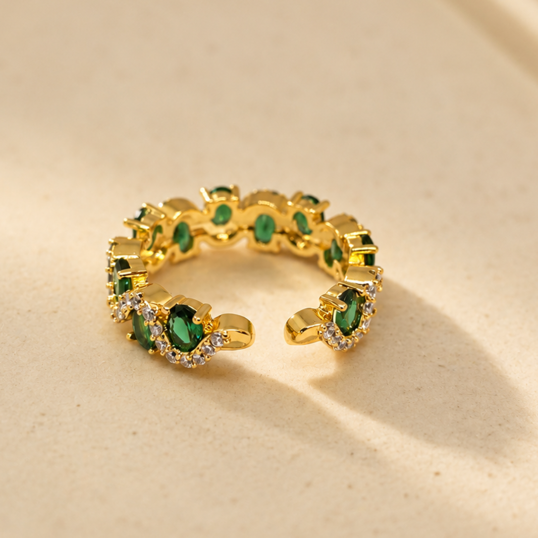 A gold open ring band decorated with small green heart-shaped stones and clear crystal accents, resting on a warm beige textured surface.