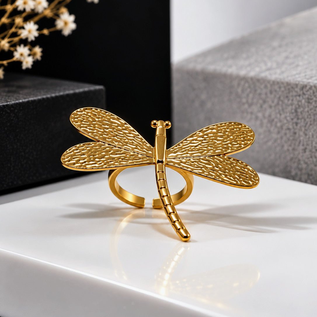 A gold dragonfly ring with textured wings and a polished band, displayed on a white surface with a black and gray background.