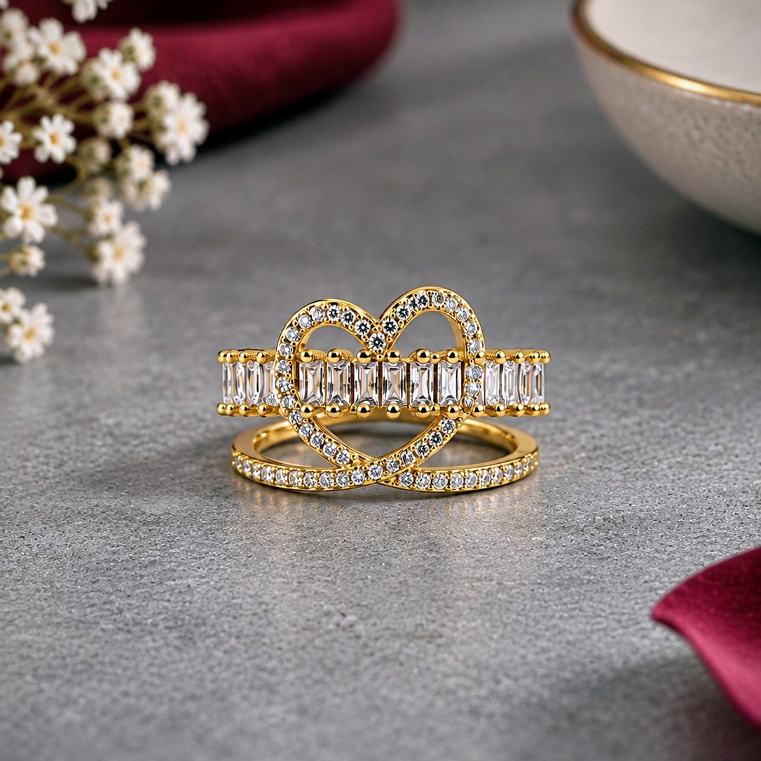 A pair of gold rings adorned with clear baguette and round-cut stones, displayed on a textured gray surface with a dark red fabric and small white flowers in the background.