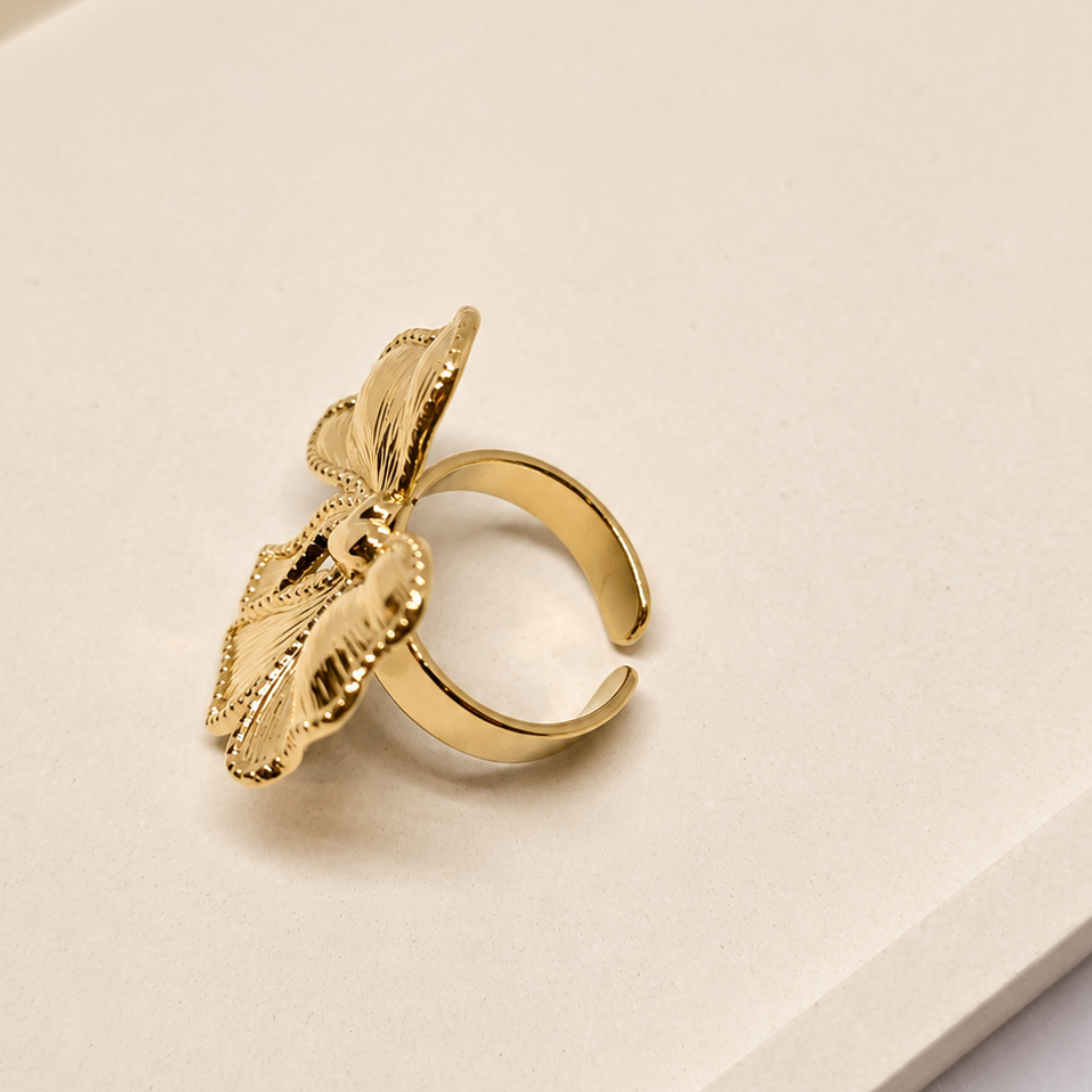 A gold-toned open ring with a sculpted leaf design, photographed on a clean cream-colored surface.