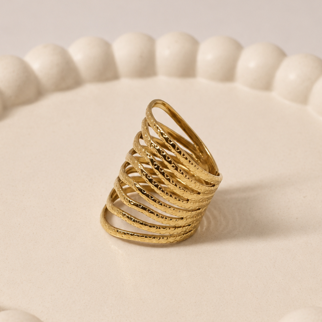 A gold-toned multi-band ring with a textured, ribbed finish, displayed on a round, cream-colored ceramic tray with a scalloped edge.