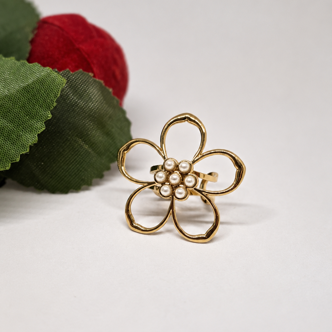 A gold flower-shaped hair claw clip with a cluster of small white faux pearls in the center, displayed on a white surface next to decorative green and red fabric leaves.