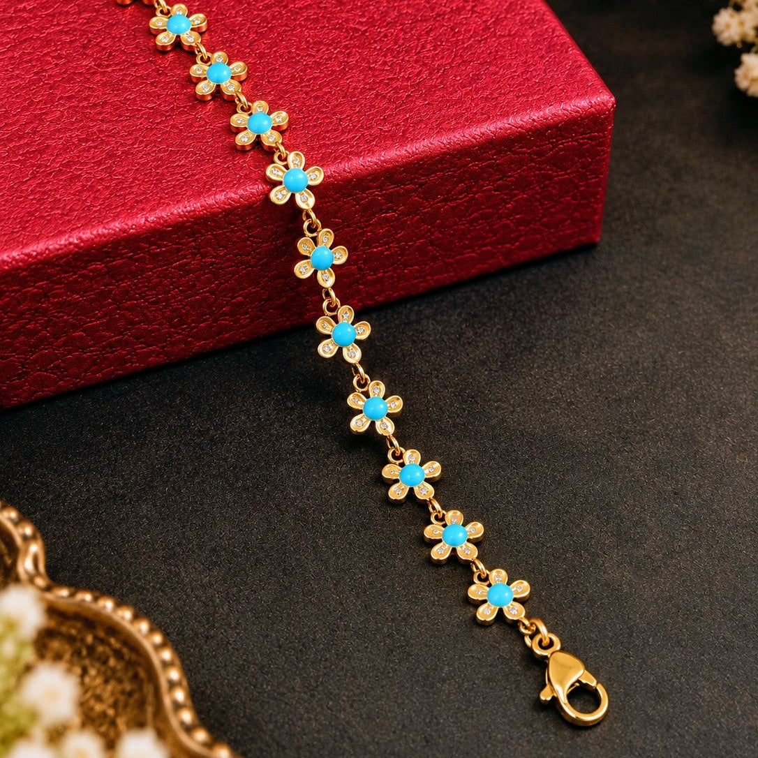 A gold chain bracelet with small flower charms featuring bright turquoise blue centers, displayed diagonally on a textured red gift box against a dark gray background with decorative floral elements.