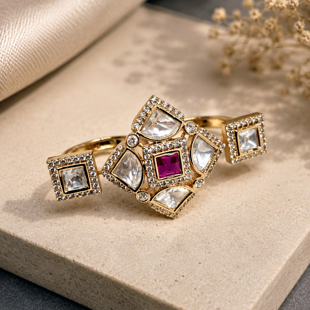 A gold-toned statement ring with clear teardrop and square stones, accented by a central deep pinkish-red square stone, displayed on a textured beige stone slab with soft cream fabric and dried flowers in the background.