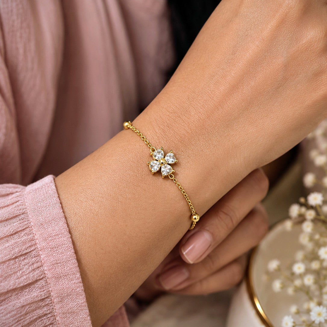 A delicate gold chain bracelet with a small four-petal flower charm set with clear stones, shown worn on a woman's wrist. The person is wearing a soft pink long-sleeve top, with a decorative bowl of baby's breath in the background.