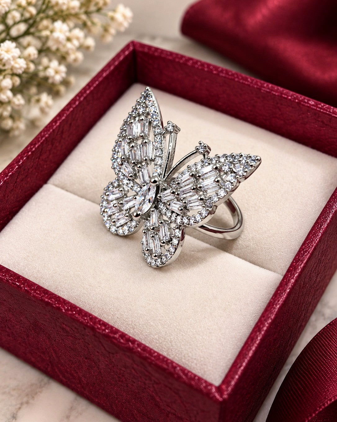 A silver butterfly ring paved with clear baguette and round-cut stones, presented inside an open red jewelry box with a cream cushion.