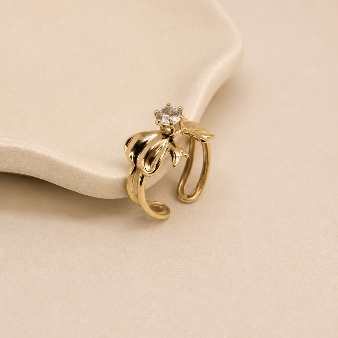 A gold-toned open ring earring with a small clear gemstone at the top, displayed on a cream-colored ceramic dish against a warm beige background.