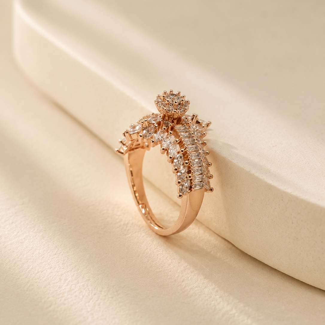 A rose gold ring with a floral crown-like top set with clear pavé stones, resting on a cream-colored textured surface.