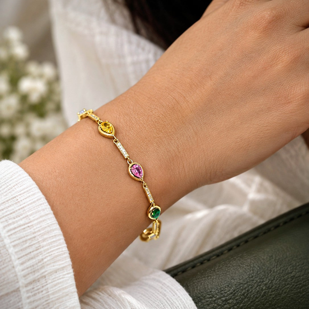 A delicate gold bracelet with colorful gemstone accents, worn on a person's wrist. The bracelet features yellow gold oval and heart-shaped charms, a pink heart charm, a green heart charm, and small clear stones.