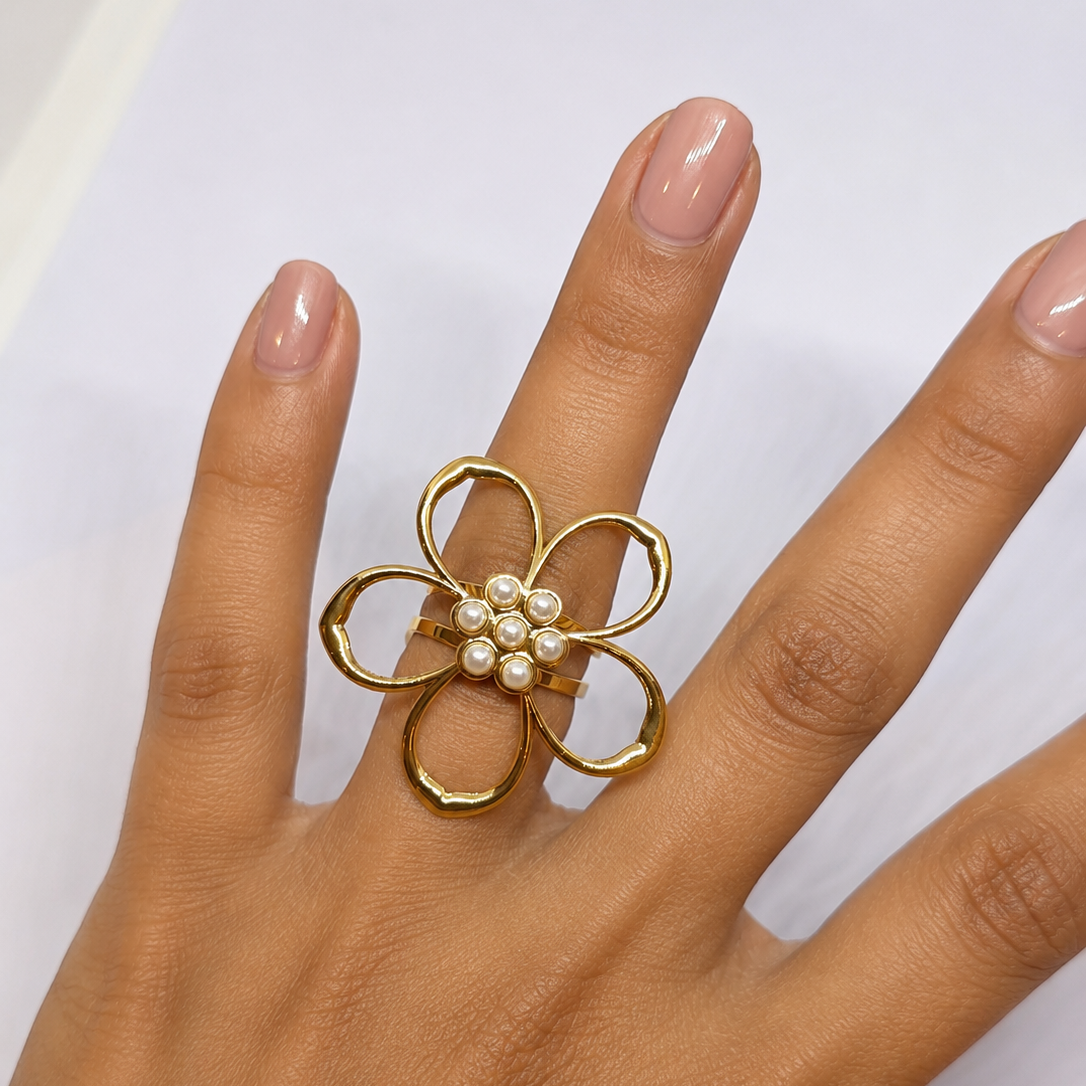 A gold flower-shaped ring with a cluster of small white pearls in the center, worn on a hand with light pink manicured nails.
