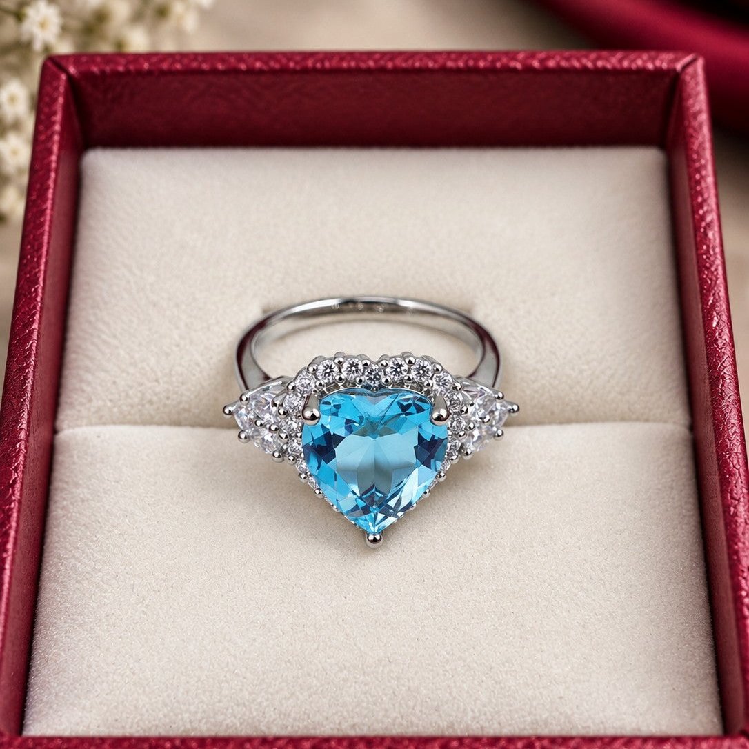 A heart-shaped light blue gemstone ring with silver-toned metal settings and small clear accent stones, displayed in an open red jewelry box with a cream insert.
