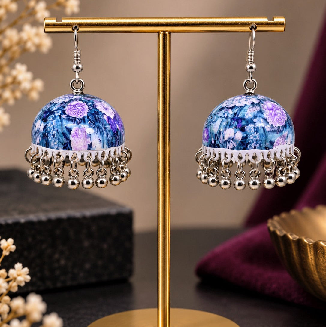 A pair of ornate jhumka earrings displayed on a gold stand. The earrings feature round blue and purple floral beads with silver dangling bell chains, set against a styled background with dried flowers and a dark surface.