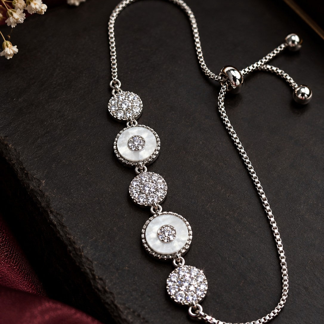 A silver-toned drop chain bracelet with five circular white and crystal-encrusted disc charms, displayed on a dark slate surface with dried flowers and burgundy fabric in the background.