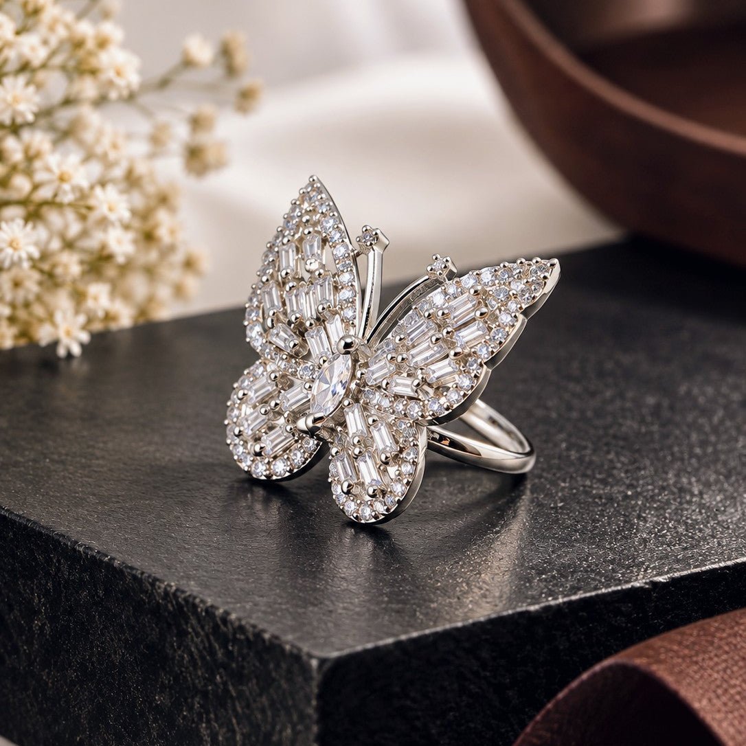 A butterfly-shaped ring set with clear baguette and round stones in a silver-toned metal, displayed on a dark textured block with soft floral and brown decorative props in the background.