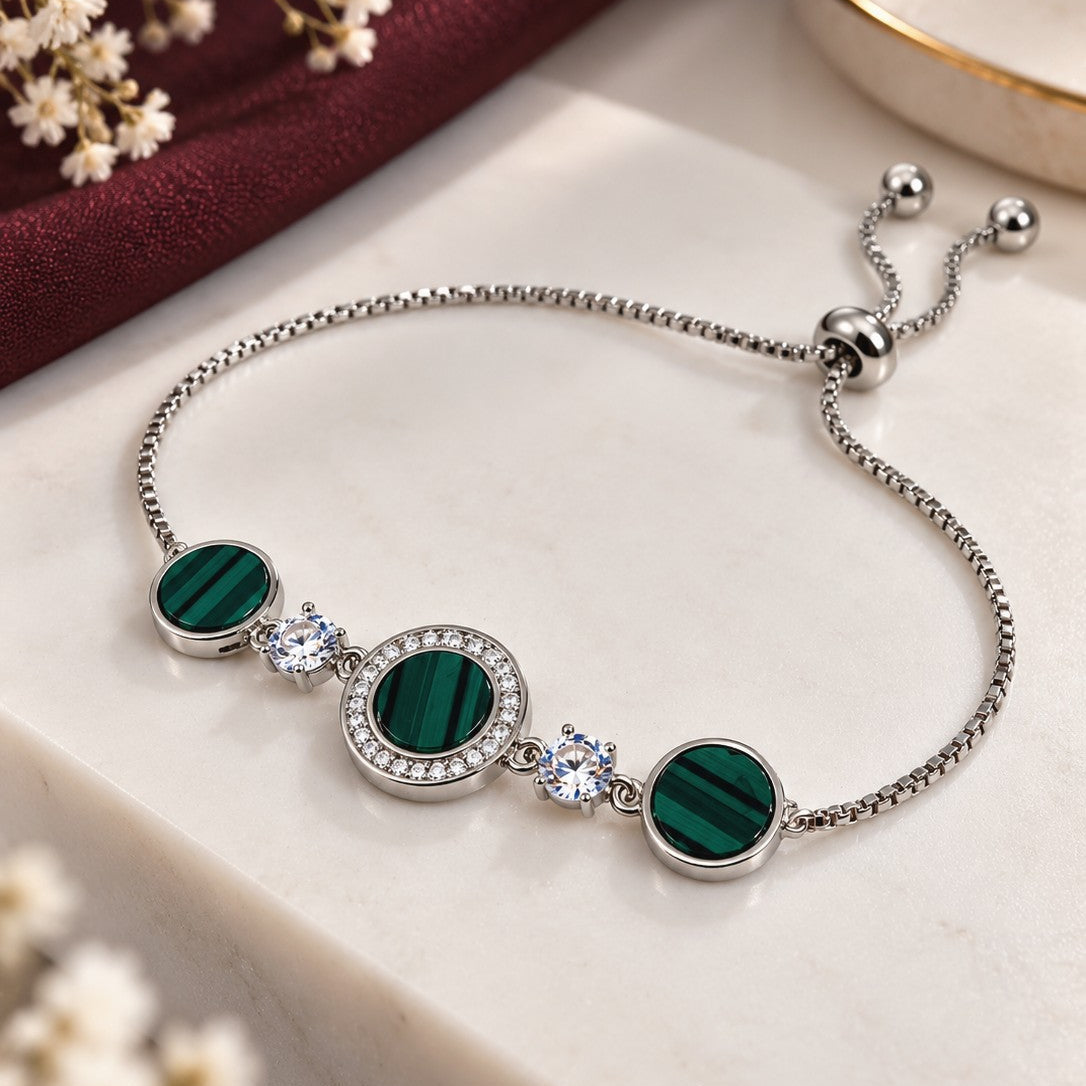 A silver-tone adjustable chain bracelet featuring three oval green malachite stones and two clear crystal accents, displayed on a cream marble block with a dark red fabric and dried flowers in the background.