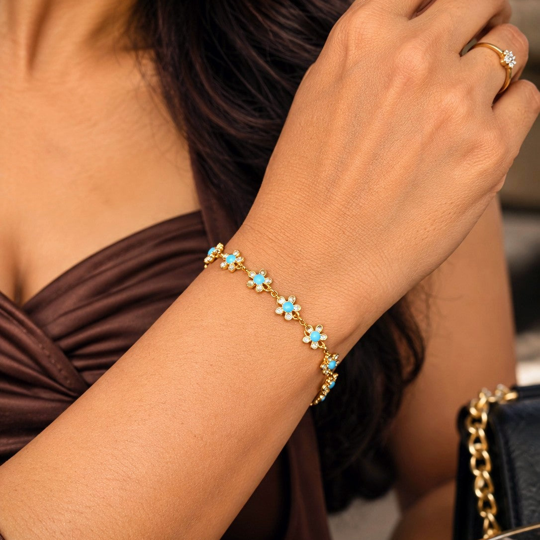 A gold-tone floral bracelet with light blue stones worn on a woman's wrist, shown in a close-up lifestyle shot. The woman is wearing a brown strapless top and a gold ring with a clear stone.