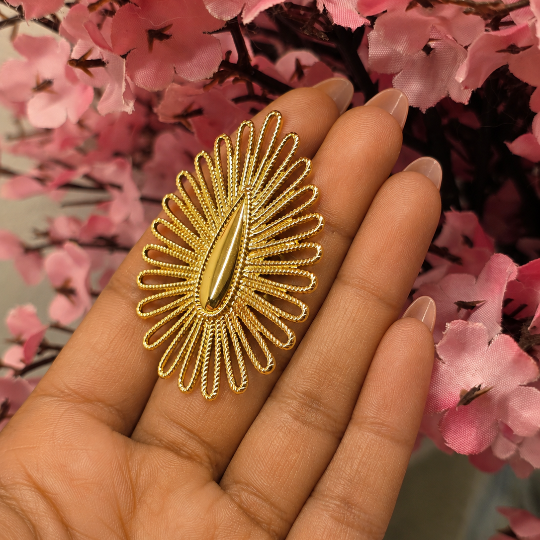 A gold-toned sunburst brooch with a teardrop center, displayed on a person's hand against a background of pink cherry blossoms.
