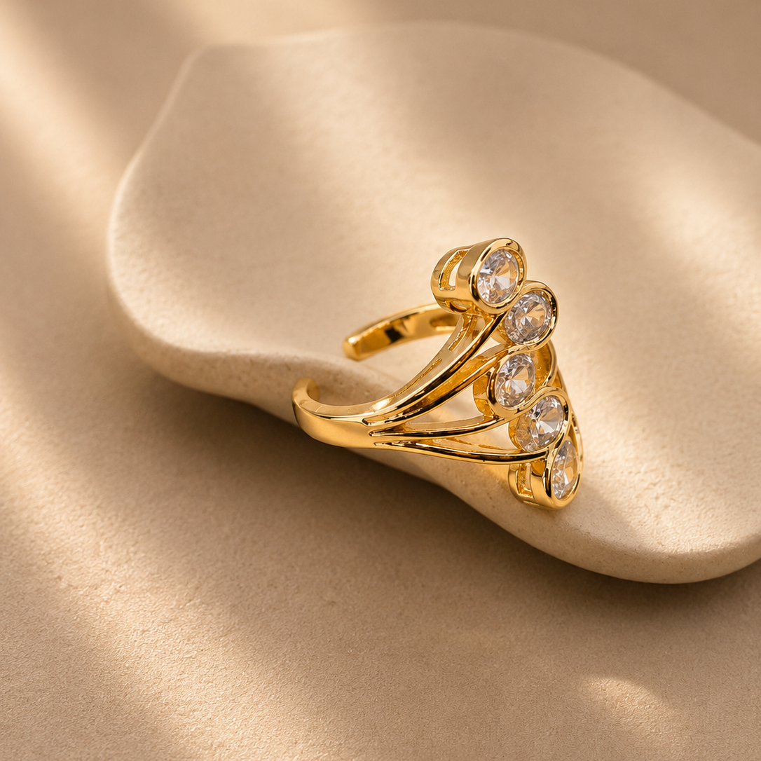 A gold ring with a cluster of clear round gemstones, displayed on a beige textured surface with soft shadows.