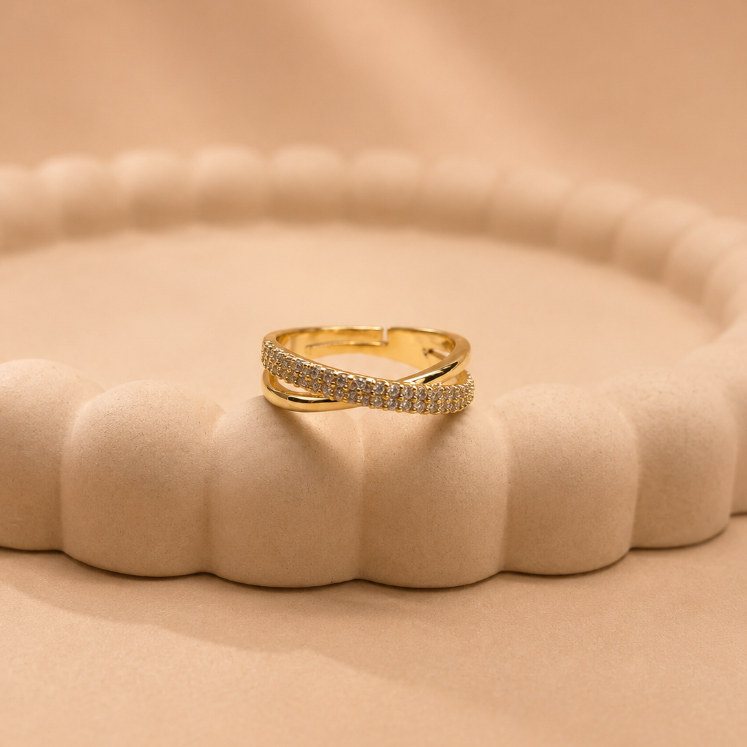 A gold ring with a pavé-set row of clear stones, displayed on a textured beige scalloped ring stand against a matching beige background.