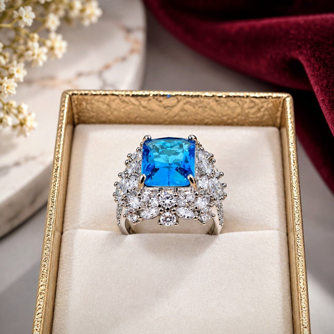 A silver-toned ring with a large square blue gemstone and surrounding clear stones, displayed in a gold jewelry box with cream lining. White flowers and a deep red fabric are softly blurred in the background.