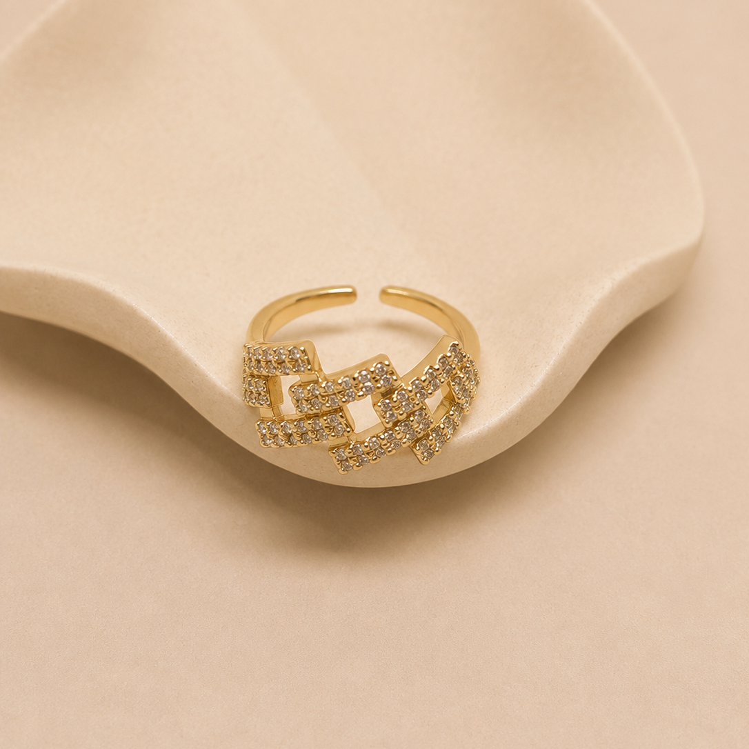 A gold-toned open ring with a geometric, interlocking chain-link design paved with small clear stones, displayed on a wavy cream ceramic dish against a warm beige background.