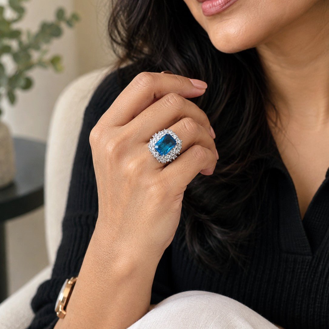 A close-up lifestyle shot of a woman wearing a statement ring with a large blue gemstone surrounded by a halo of small clear stones, set in a silver-toned metal. She is wearing a black ribbed top and white trousers, with a gold watch visible on her wrist.