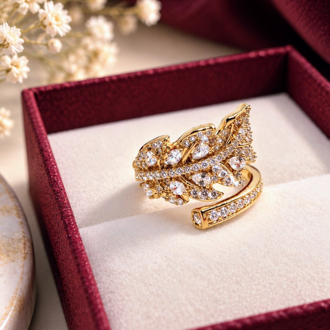 A gold ring with pavé-set clear stones in a leaf-like wrap design, displayed inside an open burgundy jewelry box with a cream insert. Small white flowers and a dark burgundy fabric are softly blurred in the background.