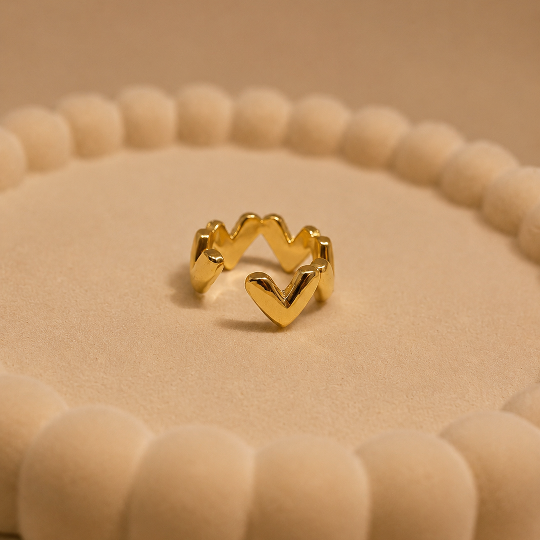 A gold heart-shaped ring displayed on a textured beige circular stand against a warm beige background.