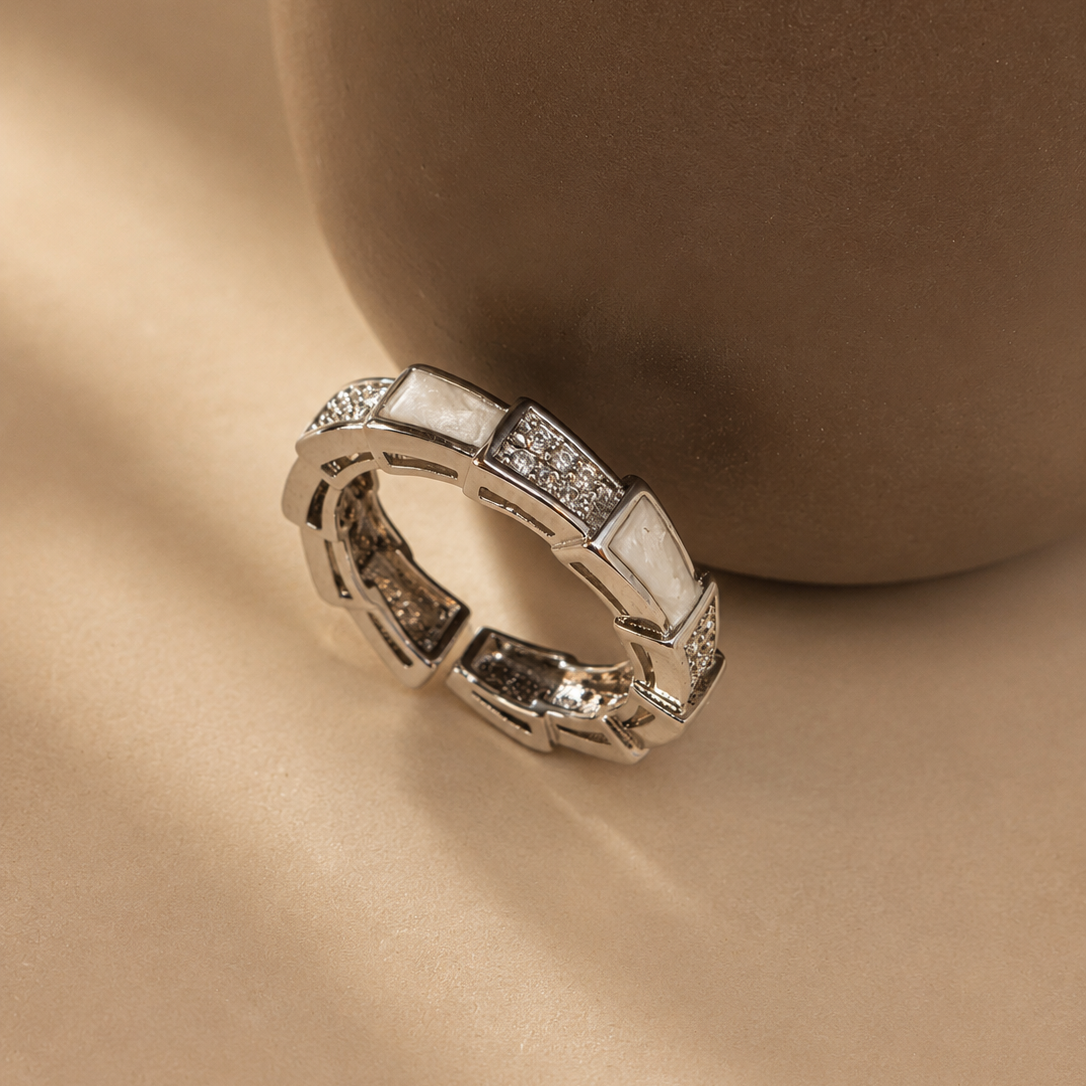 A silver-toned geometric ring with white inlay panels and small clear stones, photographed on a warm beige surface with a large brown ceramic vase in the background.