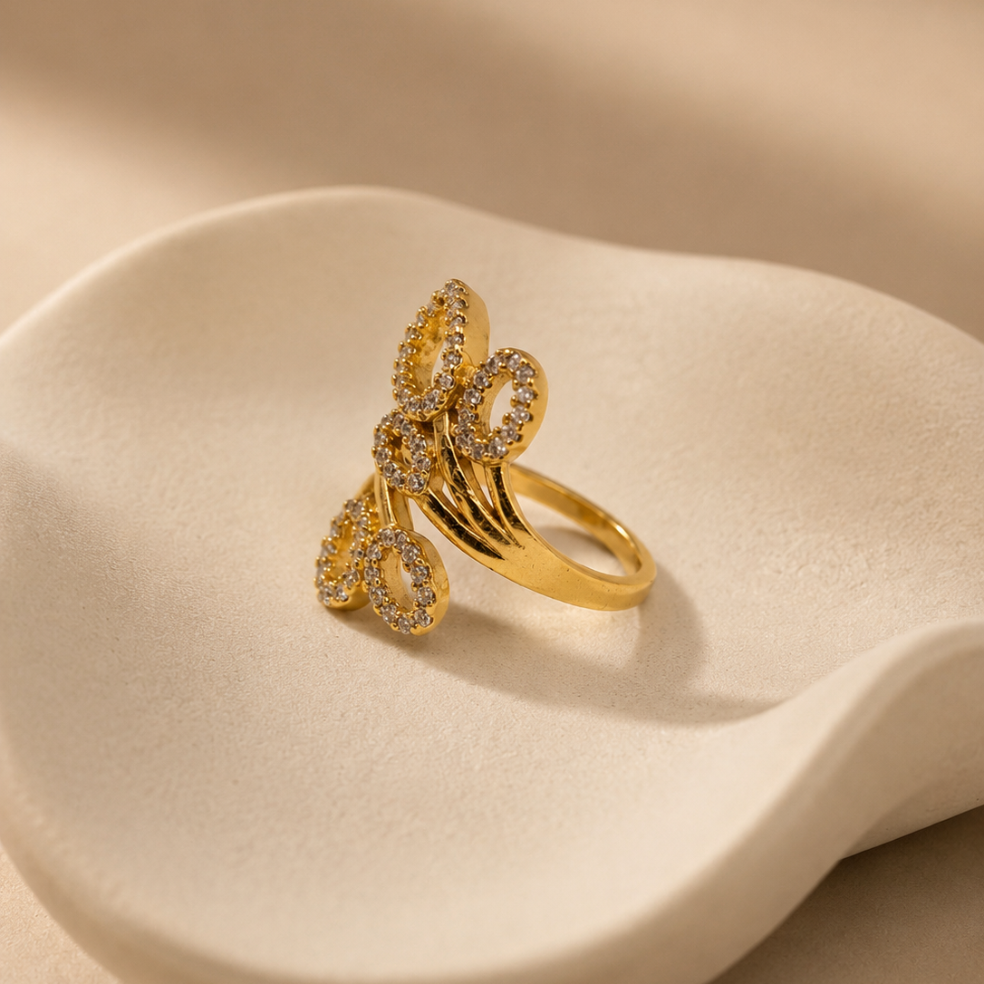 A gold ring with a sculptural, multi-looped design paved with small clear stones, displayed on a wavy, cream-colored ceramic stand against a warm beige background.