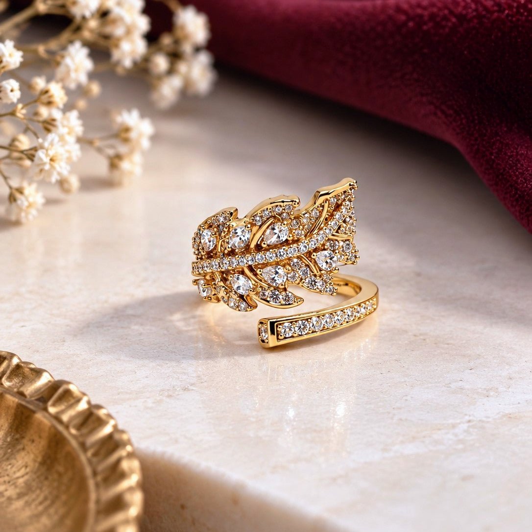A gold leaf-shaped ring embellished with clear pavé stones, displayed on a light marble surface with a deep burgundy velvet cloth and small white flowers in the background.