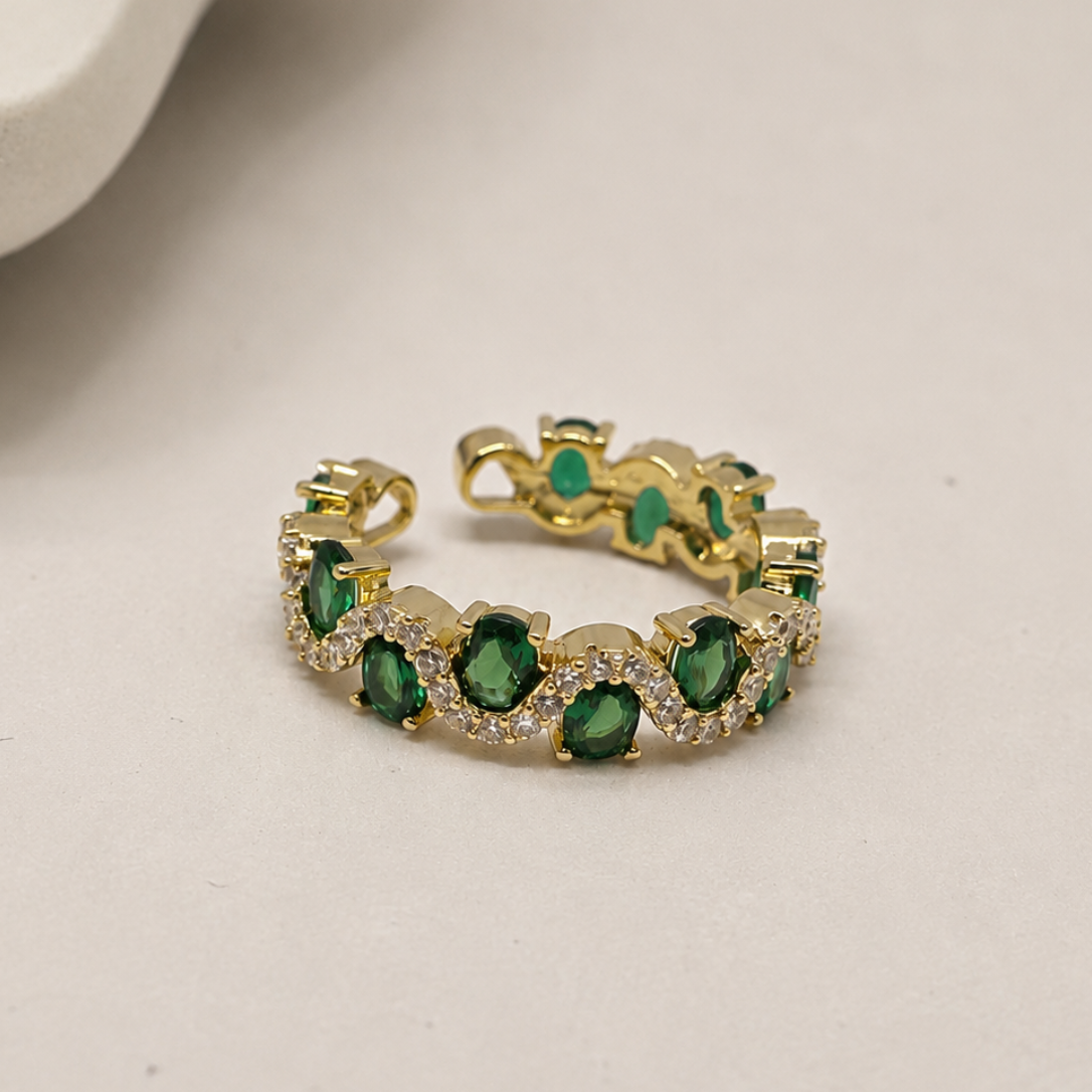 A gold-toned stretch ring featuring a row of green oval gemstones and small clear accent stones, photographed on a light neutral surface with a soft shadow.