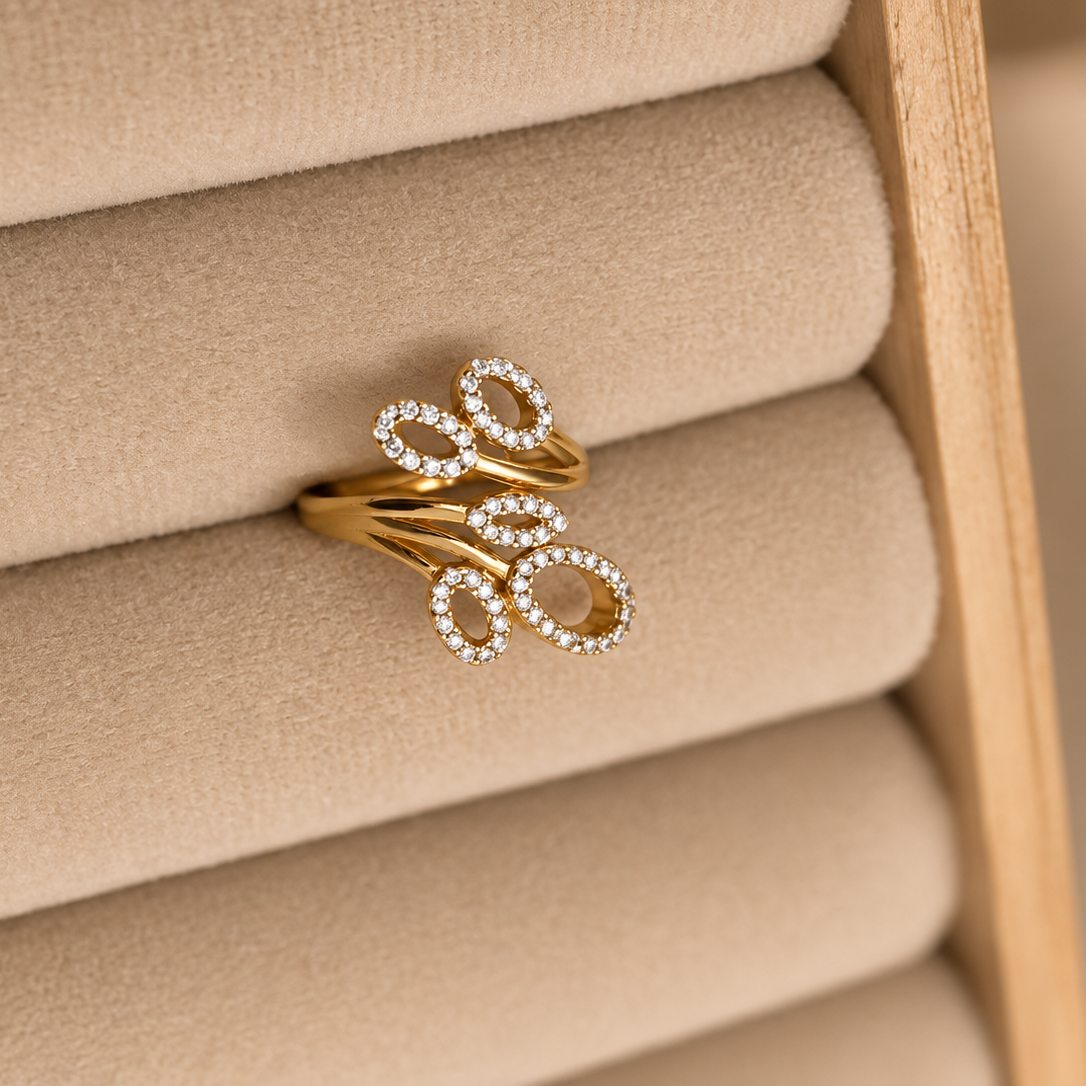 A gold ring with a stylized bow design set with small clear stones, displayed on a beige fabric ring roll inside a wooden jewelry box.