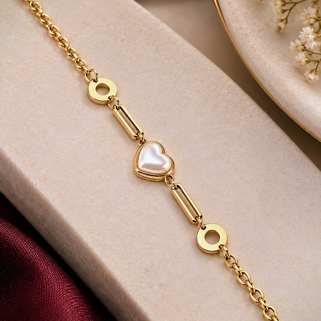 A gold chain bracelet with a central white pearl heart charm, displayed on a beige jewelry box with burgundy fabric and dried flowers in the background.